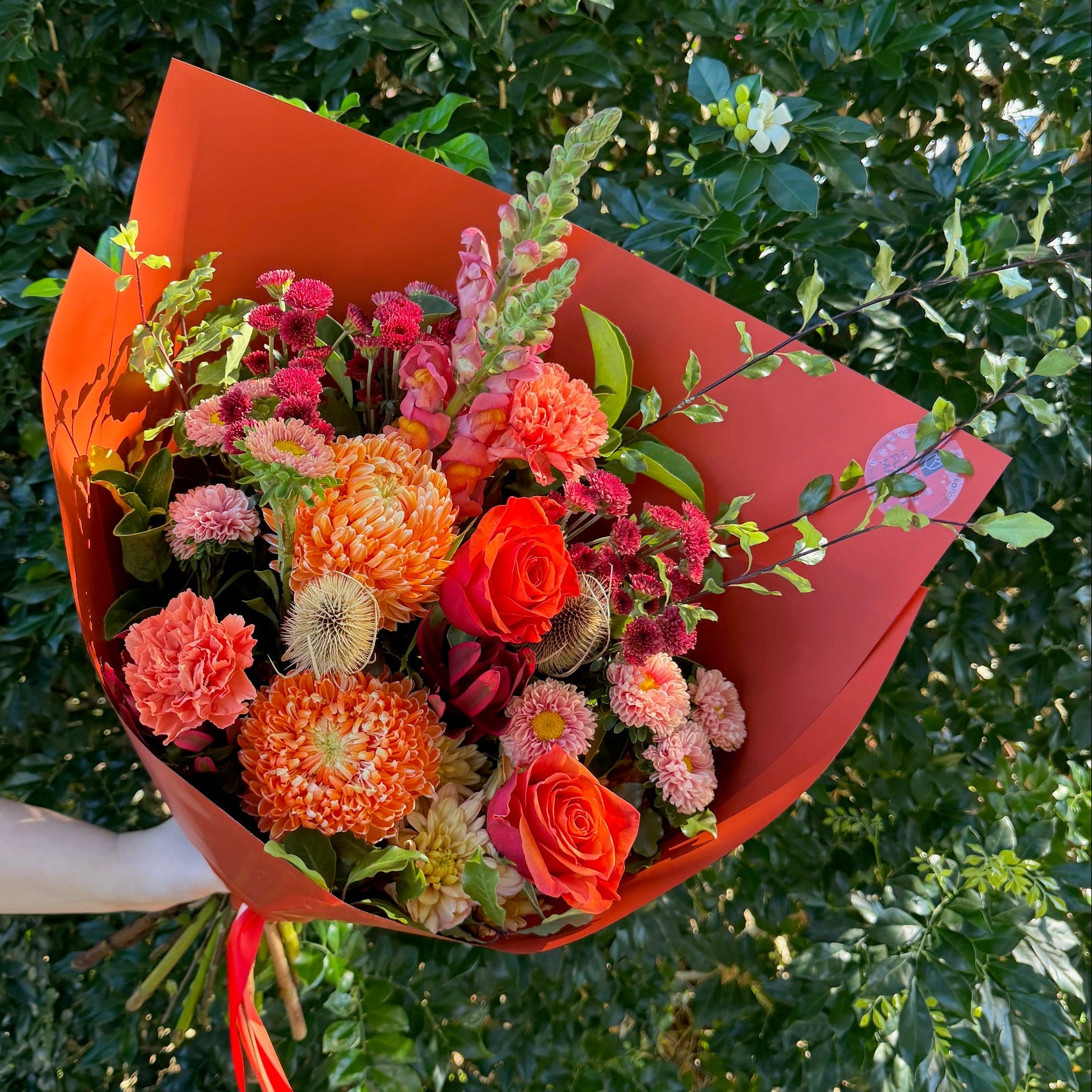 Bouquet of flowers wrapped in burnt orange paper held by a person against a green leafy background