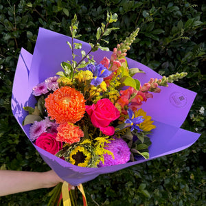 Colorful bouquet of flowers wrapped in purple paper held against a green bush background
