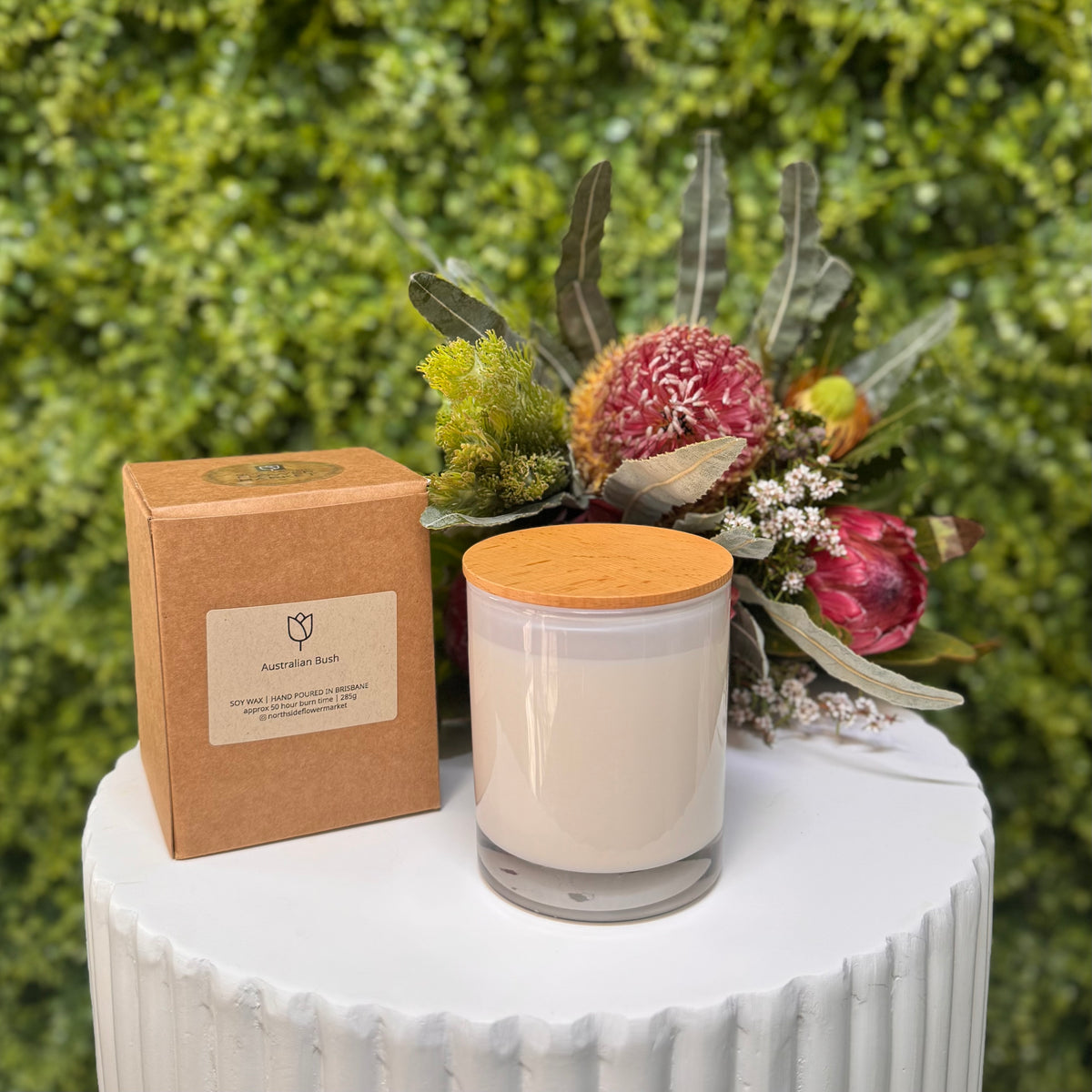 Candle with wooden lid and packaging on a white surface with a green foliage background