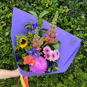 Bouquet of flowers in a purple paper wrap held against a green hedge.