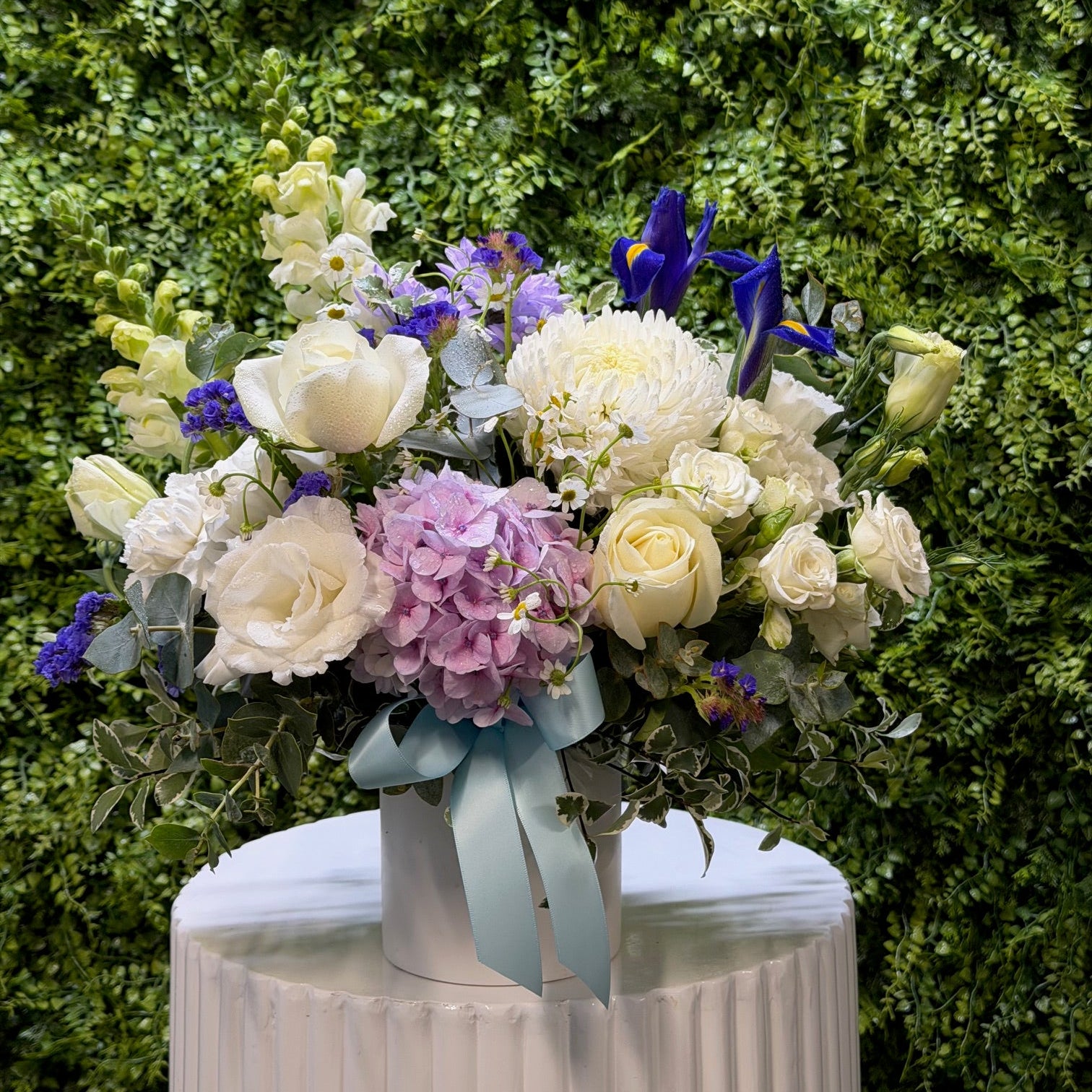Bouquet of white, pink and blue flowers with a white vase against a green hedge background