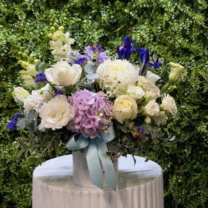 Bouquet of white, pink and blue flowers with a white vase against a green hedge background