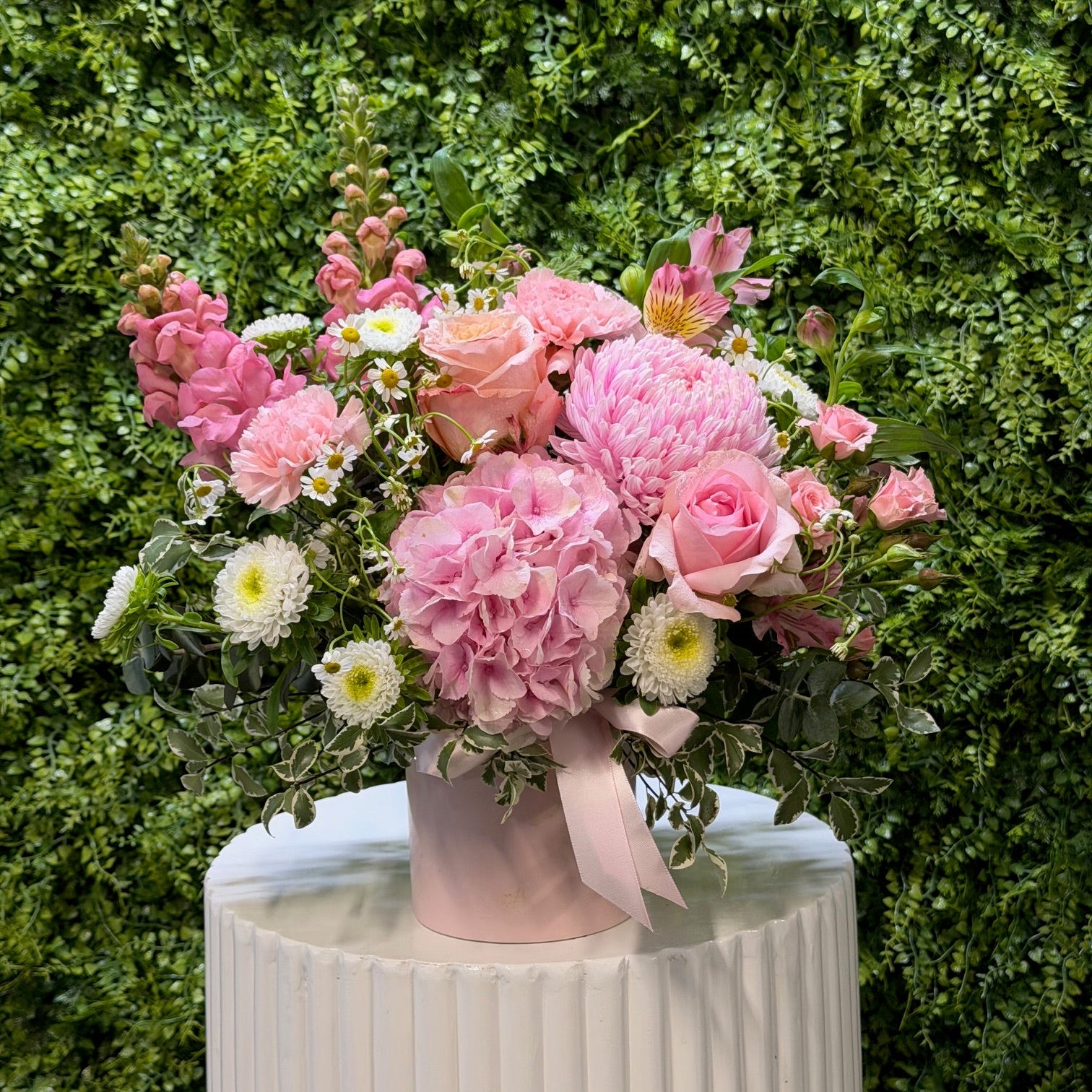 Bouquet of pink flowers with greenery on a white pedestal against a green hedge background