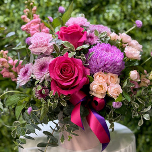 Bouquet of pink and purple flowers with a ribbon on a white pedestal against a green background