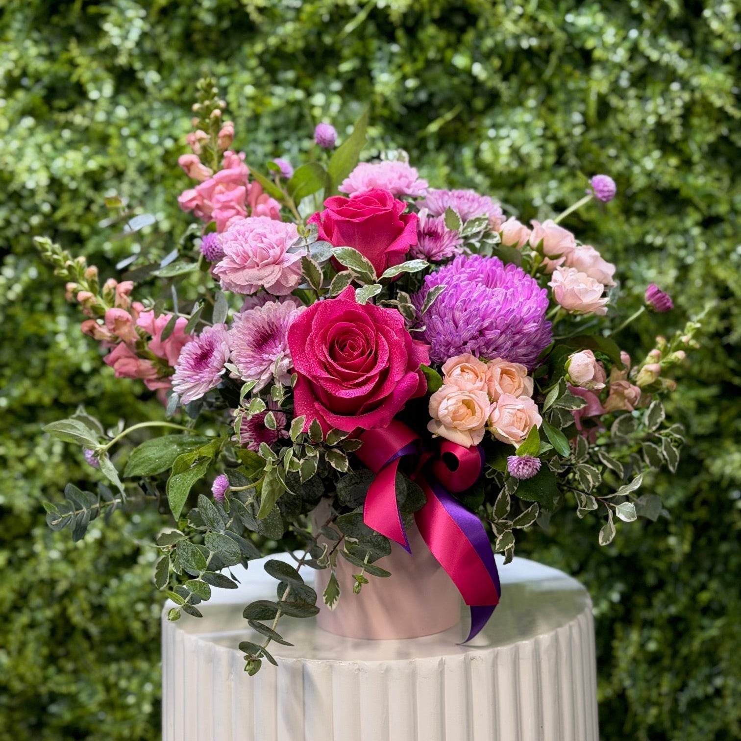 Bouquet of pink and purple flowers with a ribbon on a white pedestal against a green hedge.