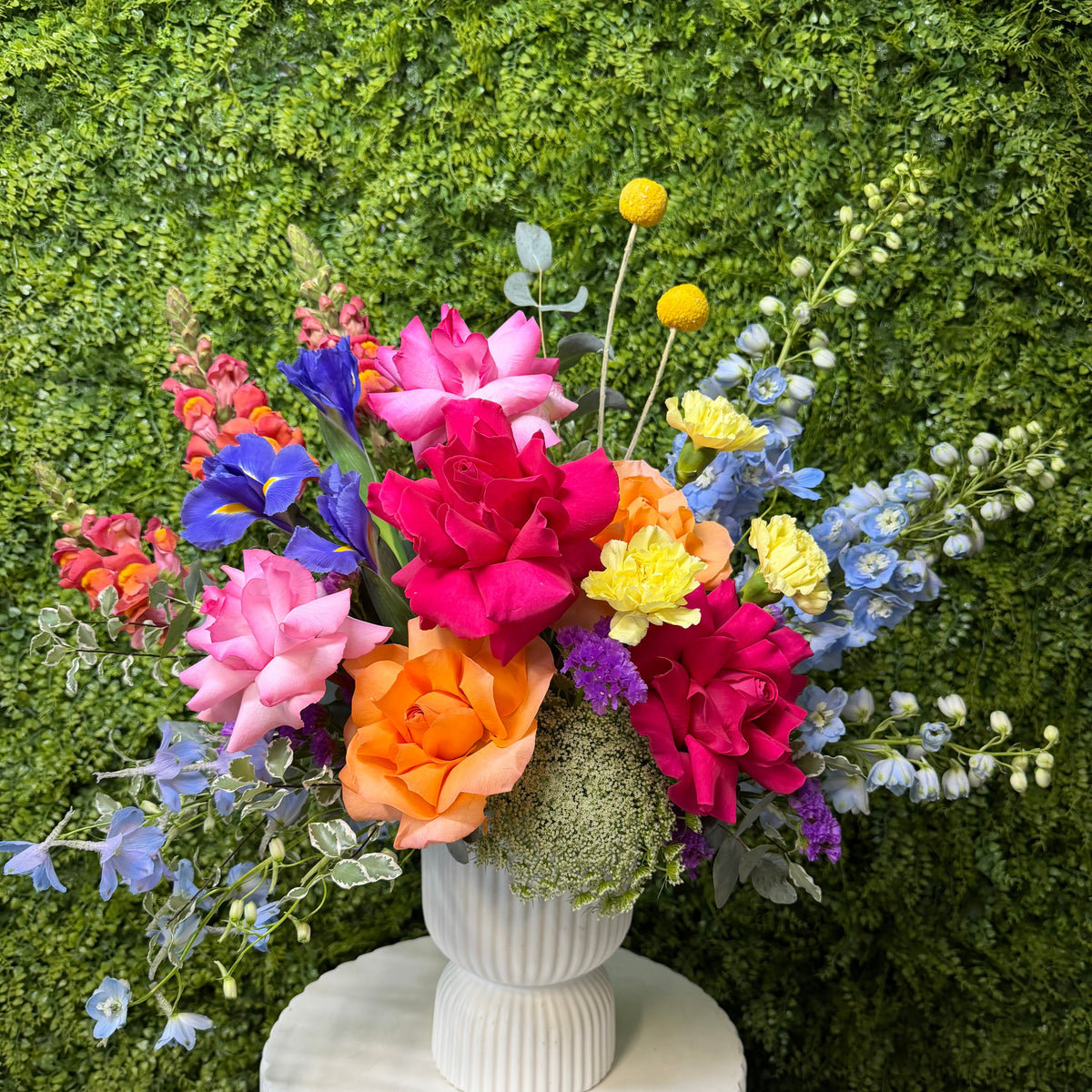 A vibrant, multicolor Spring floral arrangement featuring pink, orange, yellow, blue, and purple flowers in a ribbed white ceramic vase, displayed on a white pedestal against a green wall.