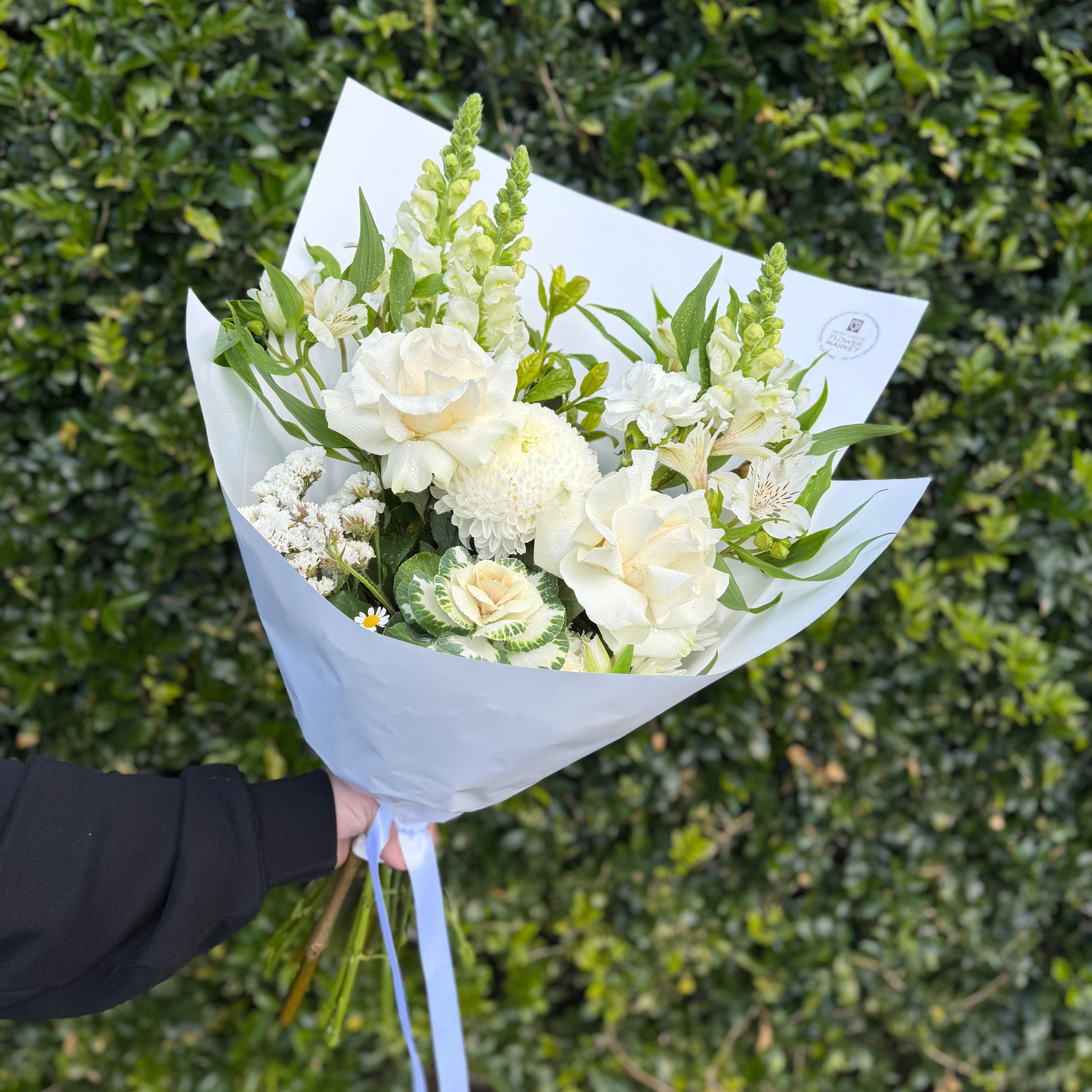 A bouquet of white flowers with green foliage, wrapped in white paper with a light blue ribbon, set against a dense green hedge background.