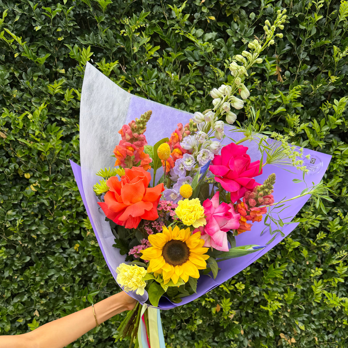 A vibrant mixed flower bouquet wrapped in light purple paper, held by a person's arm against a dense green hedge background. The bouquet features a large yellow sunflower, bright pink and orange roses, carnations, snapdragons, delphinium, and green foliage.
