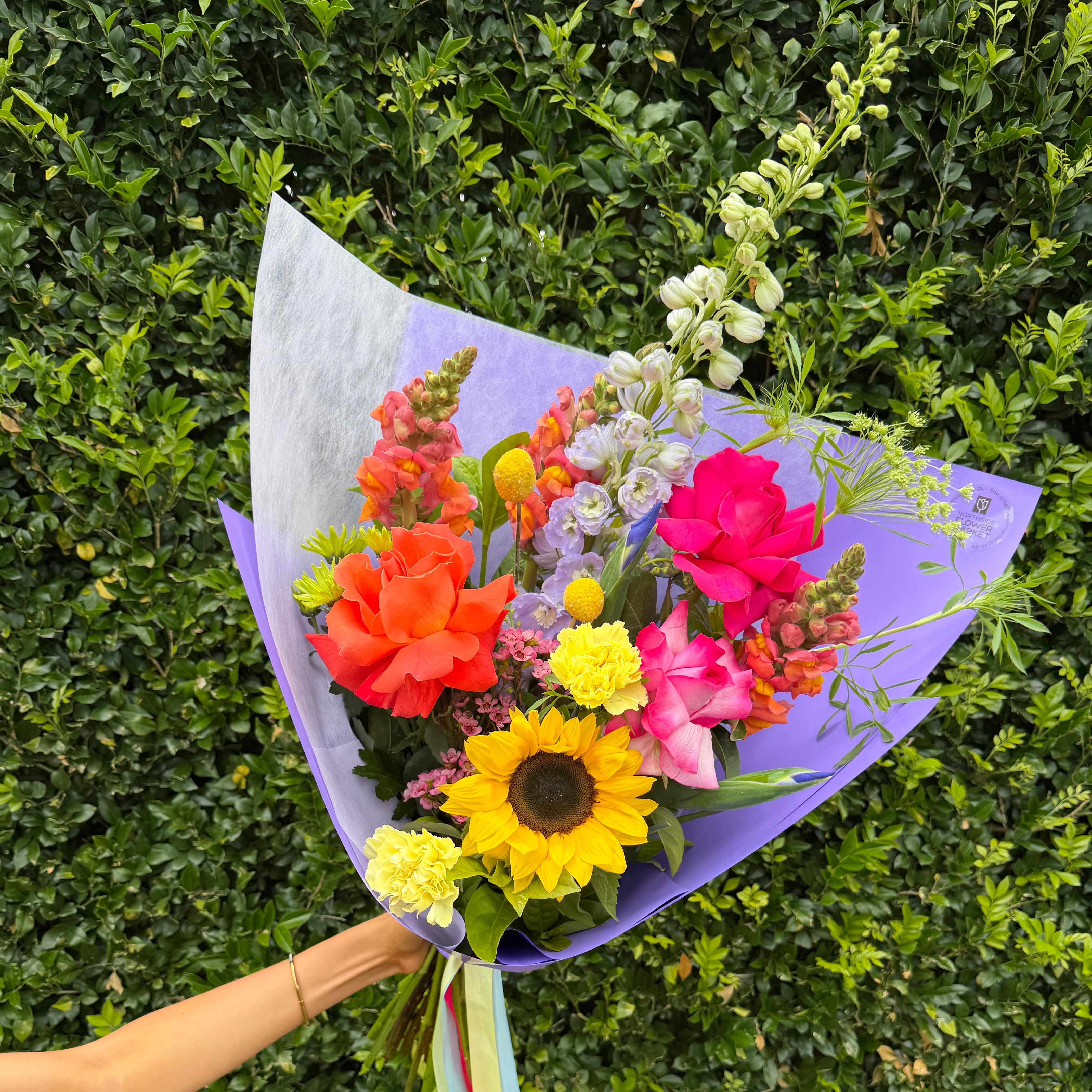 A vibrant mixed flower bouquet wrapped in light purple paper, held by a person's arm against a dense green hedge background. The bouquet features a large yellow sunflower, bright pink and orange roses, carnations, snapdragons, delphinium, and green foliage.