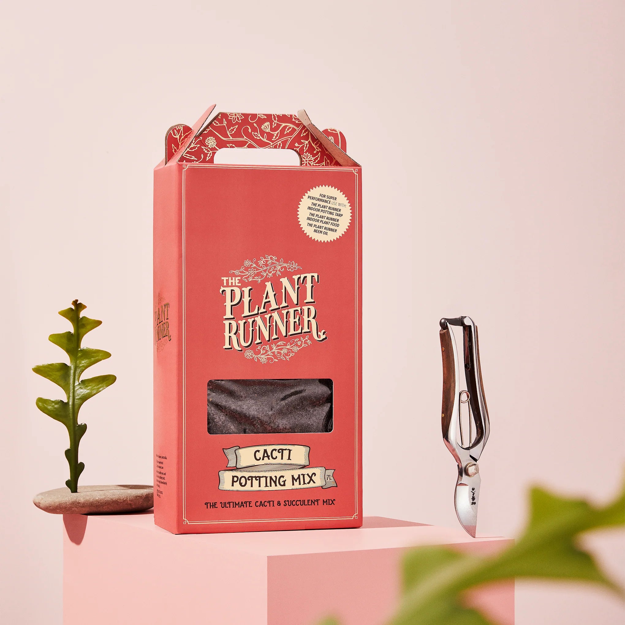 Red packaging of 'The Plant Runner' cacti potting mix with a small plant and tool on a pink background