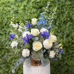 Floral arrangement with white, blue, and green flowers on a white pedestal against a green hedge background