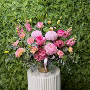 Bouquet of pink and green flowers on a white pedestal against a green hedge background