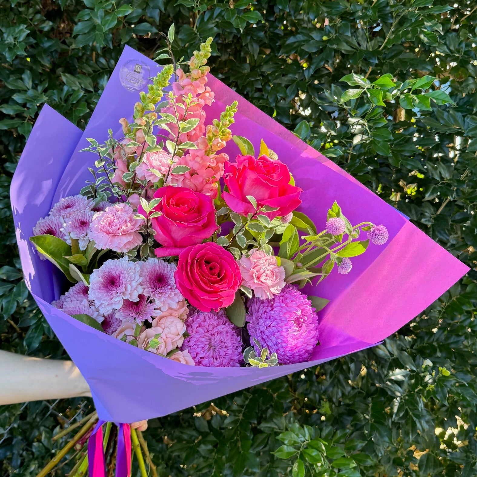Bouquet of flowers wrapped in purple paper held by a person against a green bush background