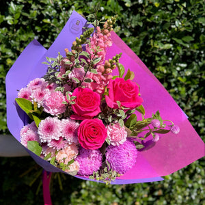 Bouquet of pink and purple flowers wrapped in purple paper with a pink ribbon against a green hedge background.