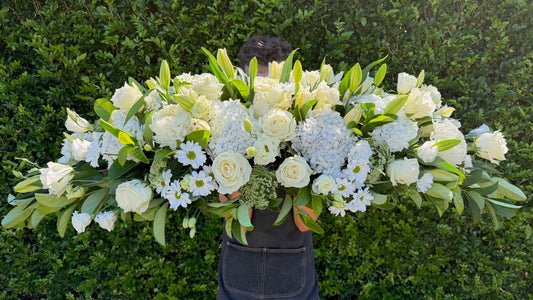Person holding a large white funeral casket spray against a green hedge.