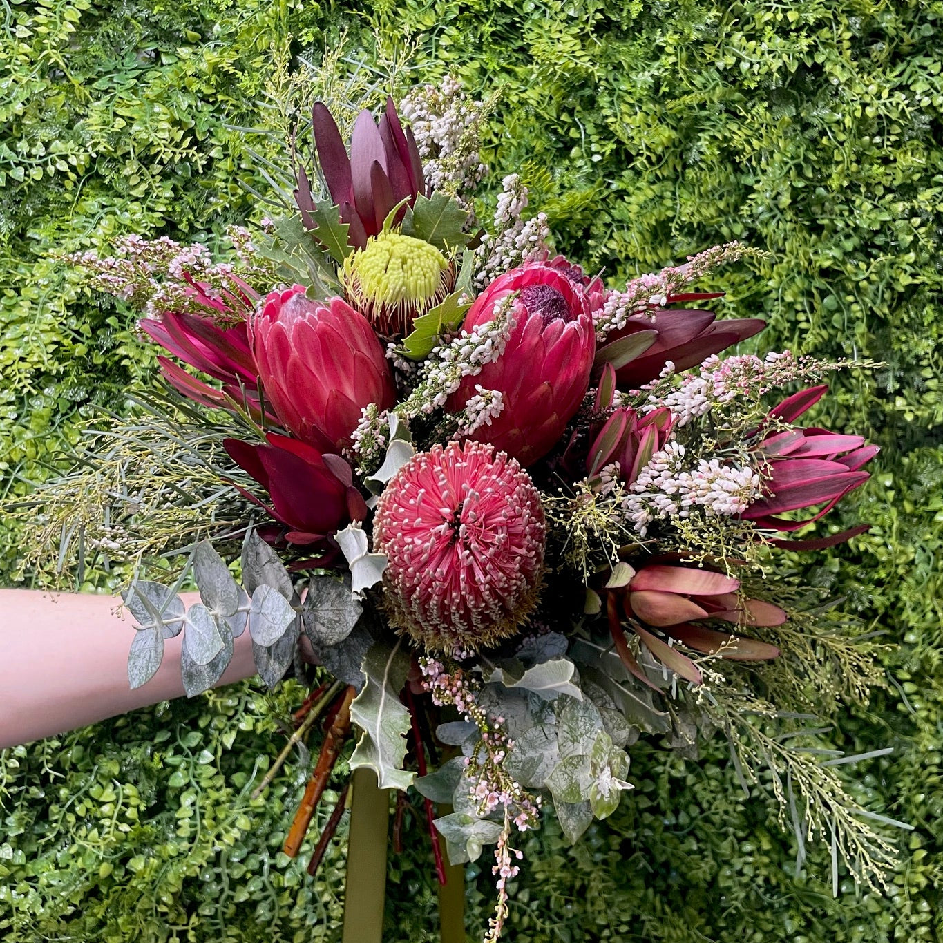 Native wedding bouquet of red proteas, banksias and leucadendron, with green foliage held against a green bush background