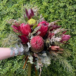 Native wedding bouquet of red proteas, banksias and leucadendron, with green foliage held against a green bush background
