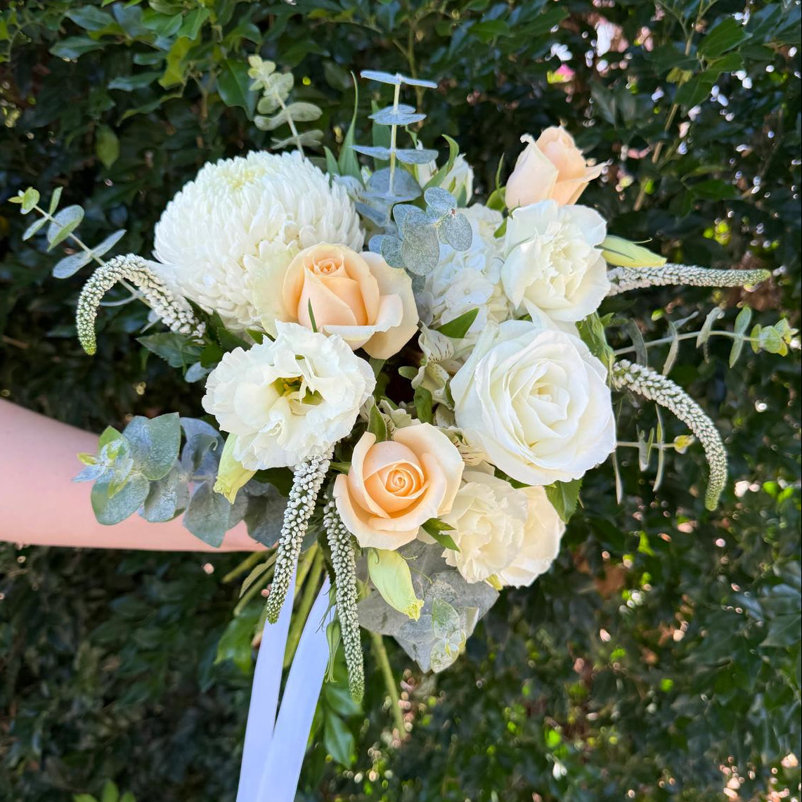 Wedding bouquet of white and peach roses with greenery held against a leafy background