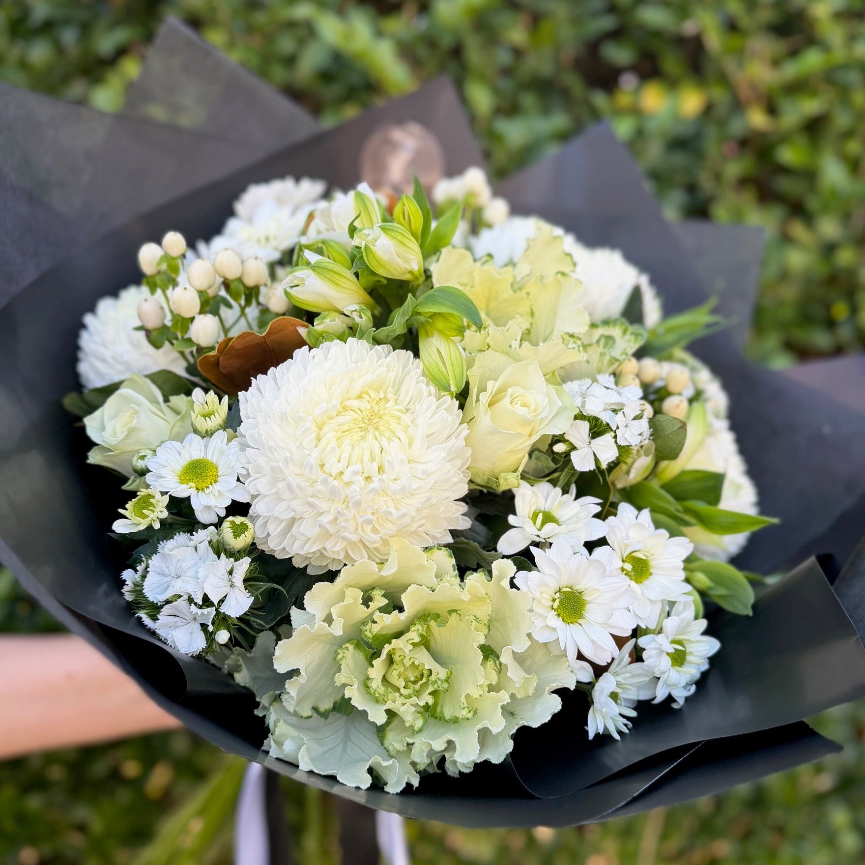 Bouquet of white and green flowers wrapped in black paper held by a person against a blurred green background.